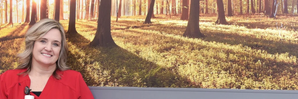 A nurse in red scrubs stands in front of a photo of trees, inside a doctor's office.