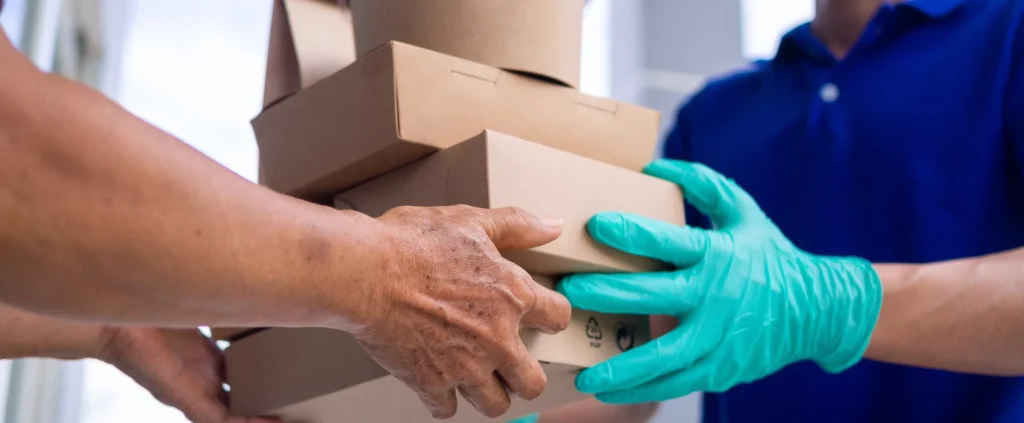 A delivery person wearing a blue shirt delivers boxes of prepared meals to a woman at home.