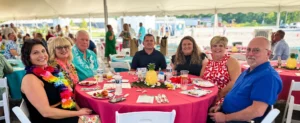 Seven people dressed in floral summer garments sits at an oval table outside, under a tent. The table has a bright red table cloth on it and is laden with beverages and games.