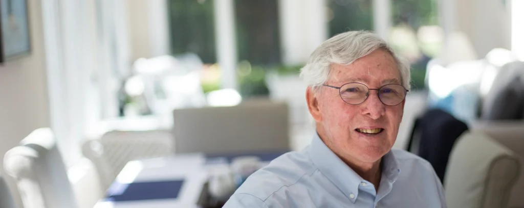 An elderly gentleman with glasses smiles while standing in his sunlit dining room.