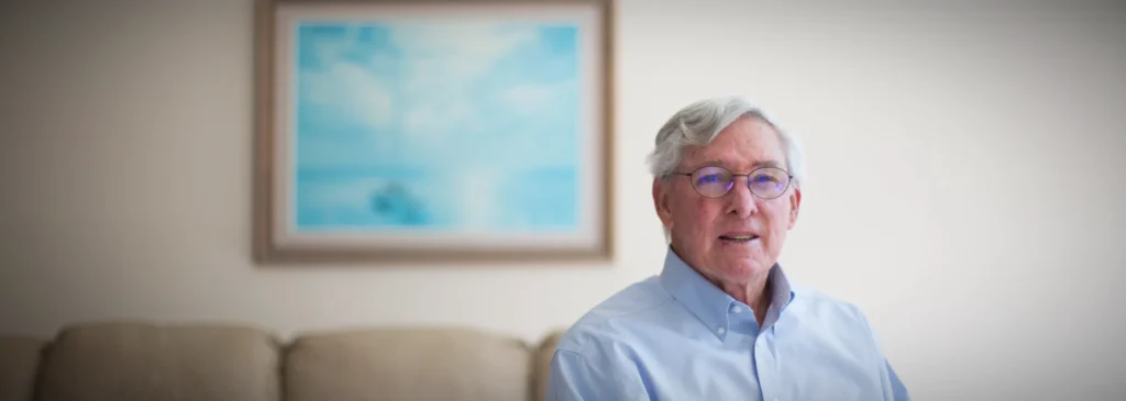 An elderly gentleman with glasses smiles while standing in his sunlit living room.