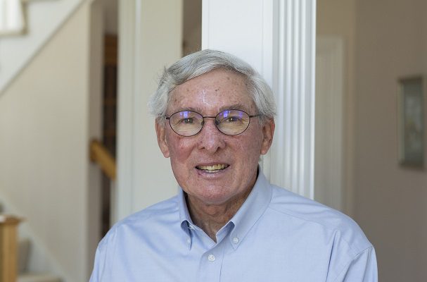 An elderly gentleman with glasses smiles while standing in his sunlit dining room.