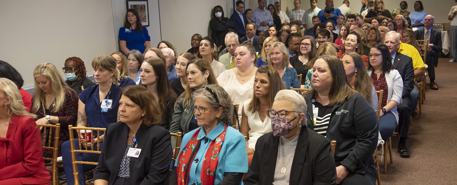 A crowd is seated in a meeting room, listening to a speaker.