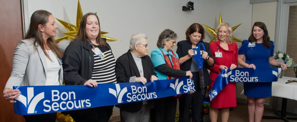 A line of women stand ready to cut a big, blue, ceremonial ribbon with a giant pair of scissors.