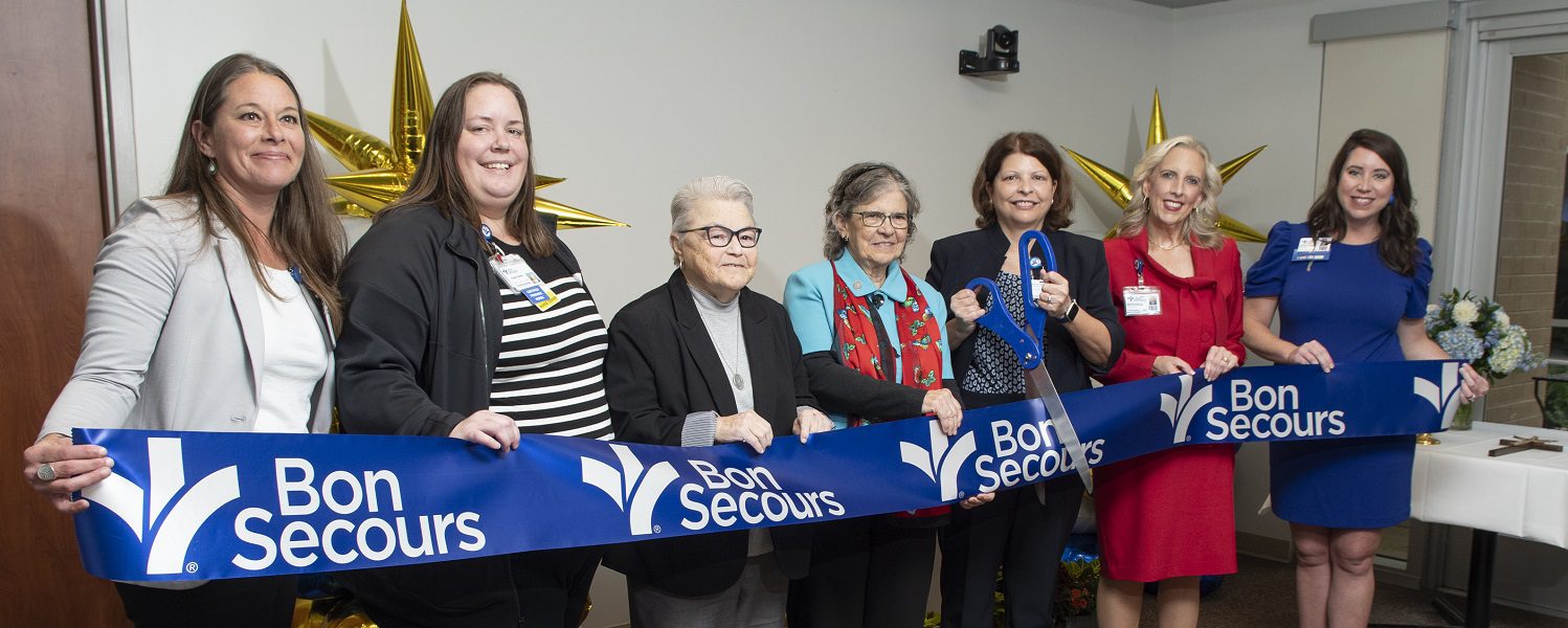 A line of women stand ready to cut a big, blue, ceremonial ribbon with a giant pair of scissors.