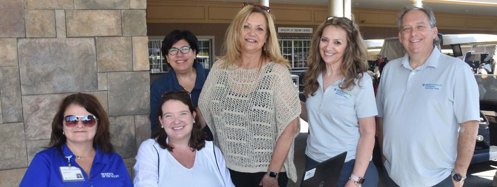 A group of smiling, professional adults pose for a picture outside on a sunny day.