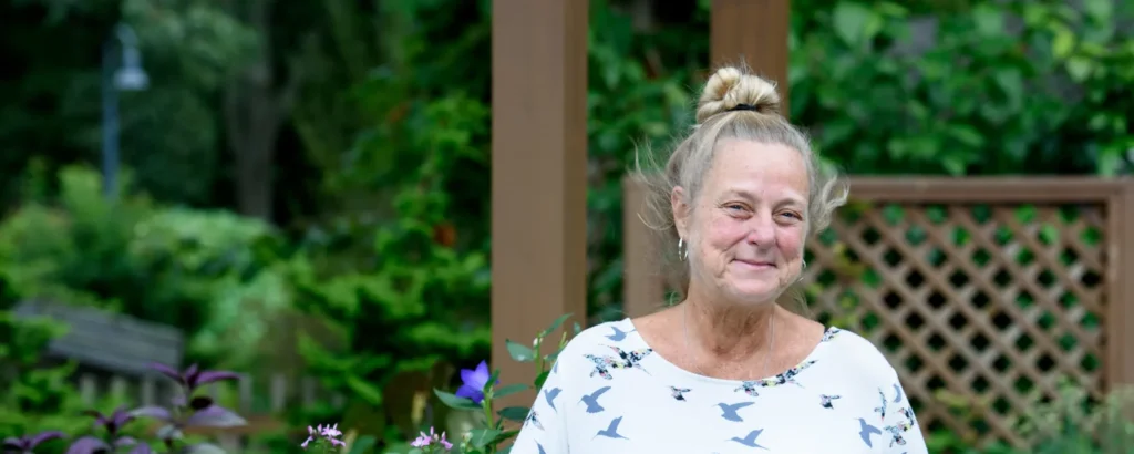 An older woman smiles while standing outside in a garden, in front of a wooden trellis. Her hair is long and blonde, and styled up in a pony tail.