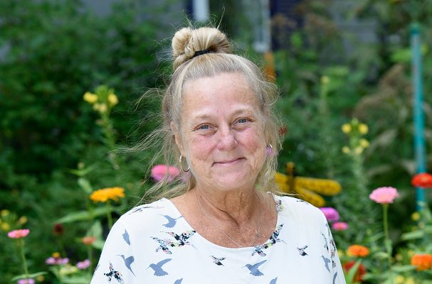 An older woman smiles while standing outside in a garden, in front of a colorful flowers. Her hair is long and blonde, and styled up in a pony tail.