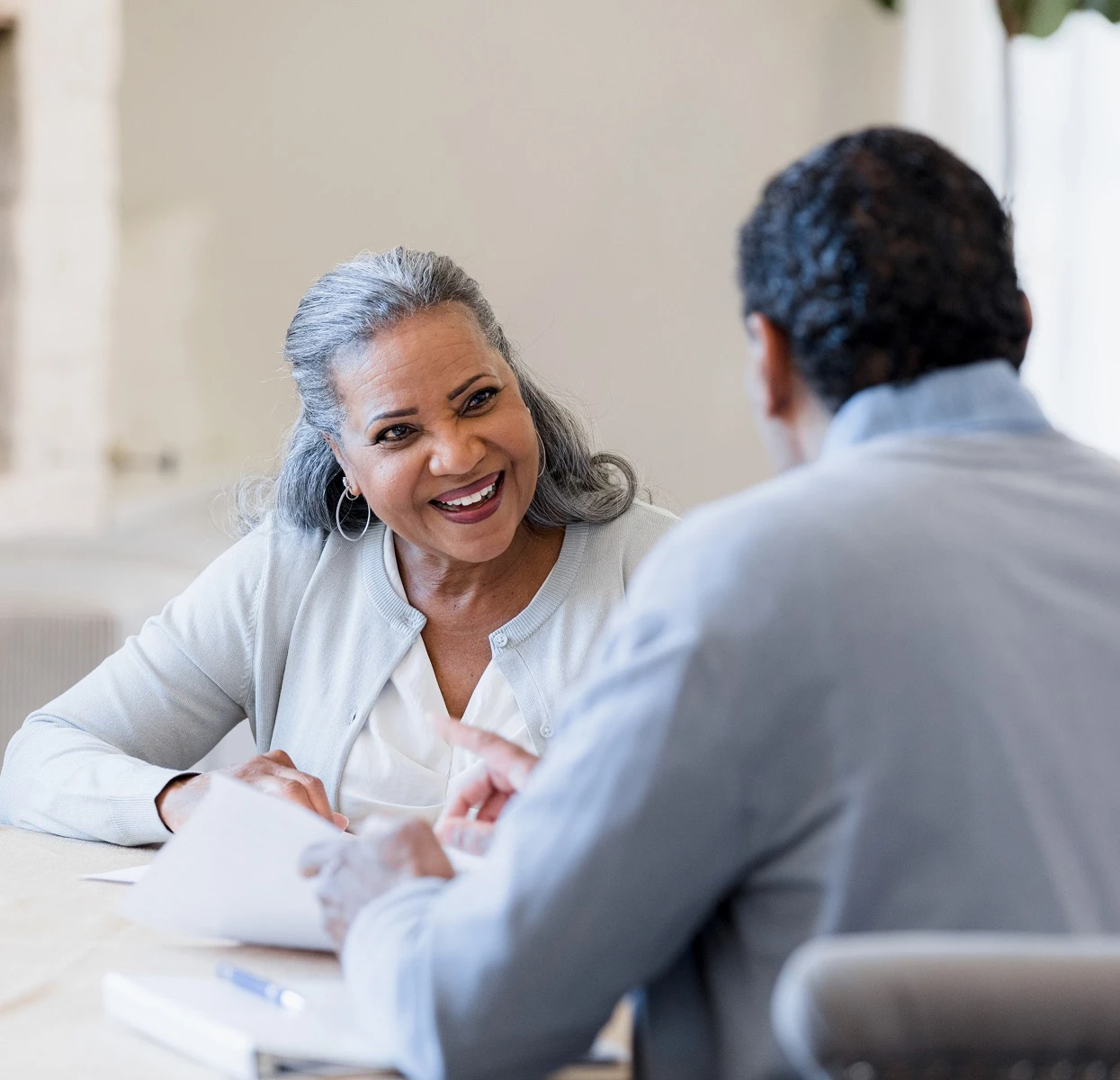 An older woman smiles brightly at a businessman, who she is sitting across the table from. There are documents on the table in between them.