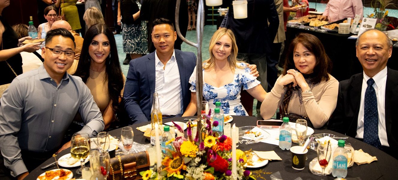 A group of friends are seated together at a formal dinner, smiling at the camera.