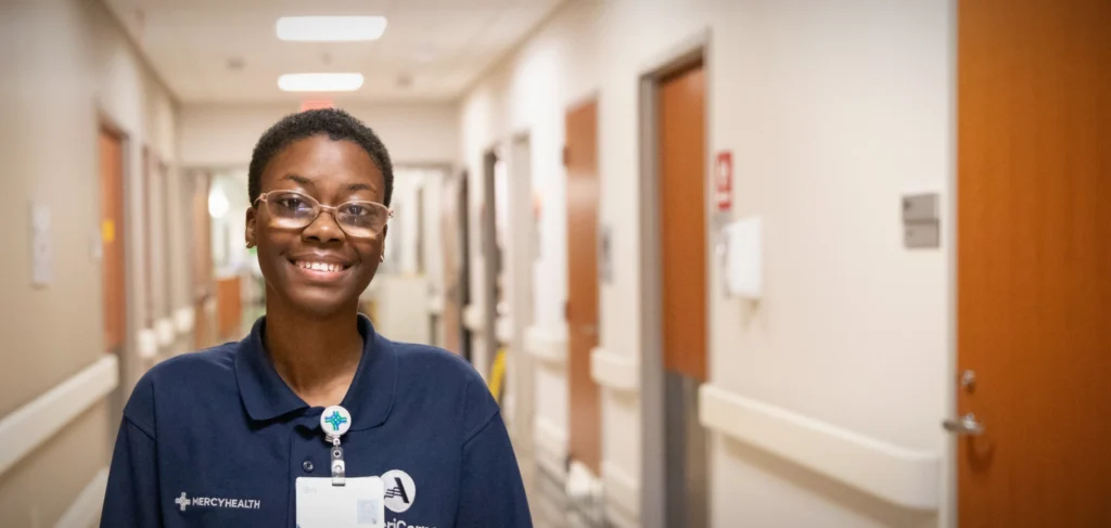 A young woman stands in a clean hospital hallway. She's wearing a navy shirt with a Mercy Health logo on it.