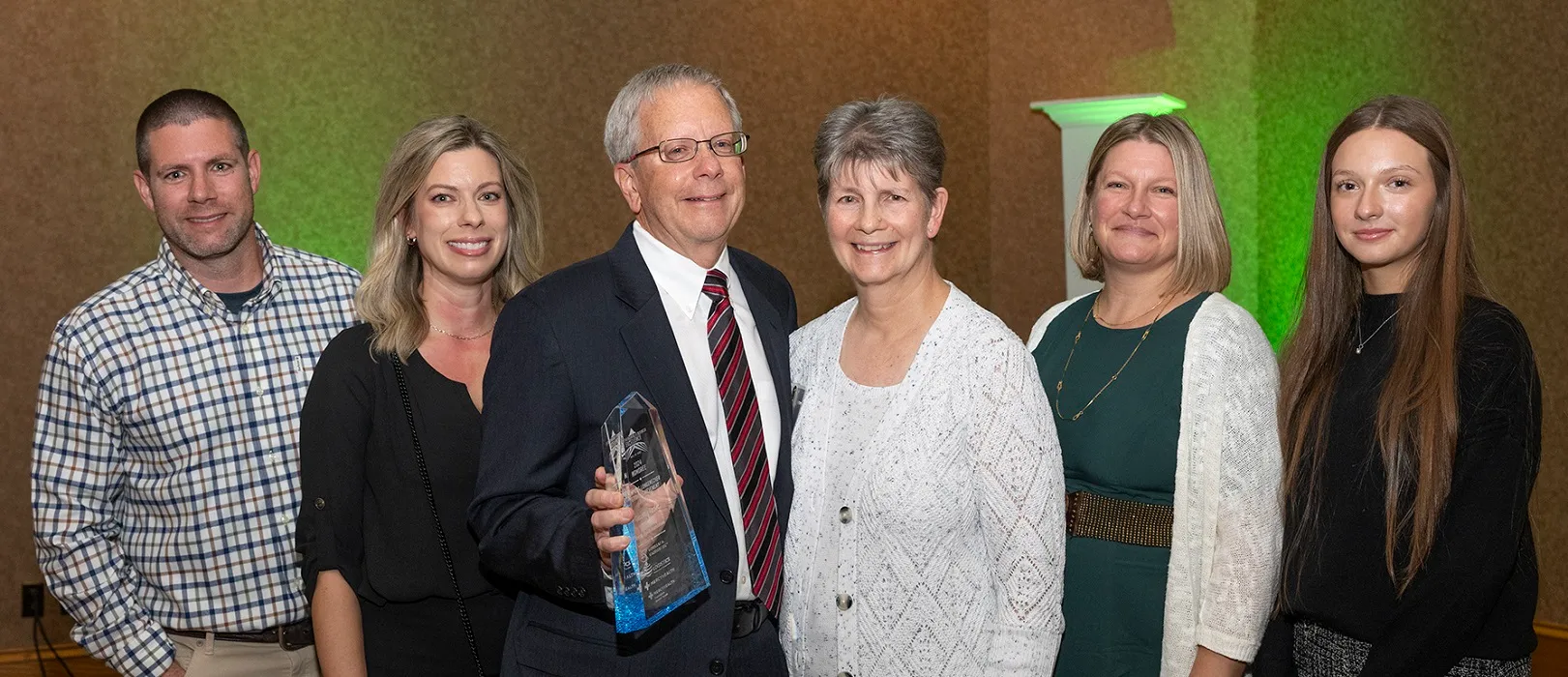 An older businessman poses for a photo with his family while holding an award.