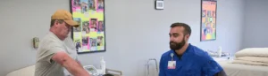 An older man in a baseball cap, t-shirt, and cargo pants stretches on a table at a physical therapy office, under the supervision of a doctor.
