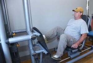 An older man in a baseball cap, t-shirt, and cargo pants exercises on a leg press.