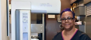 A middle-aged woman in black scrubs stands beside a mammogram machine.