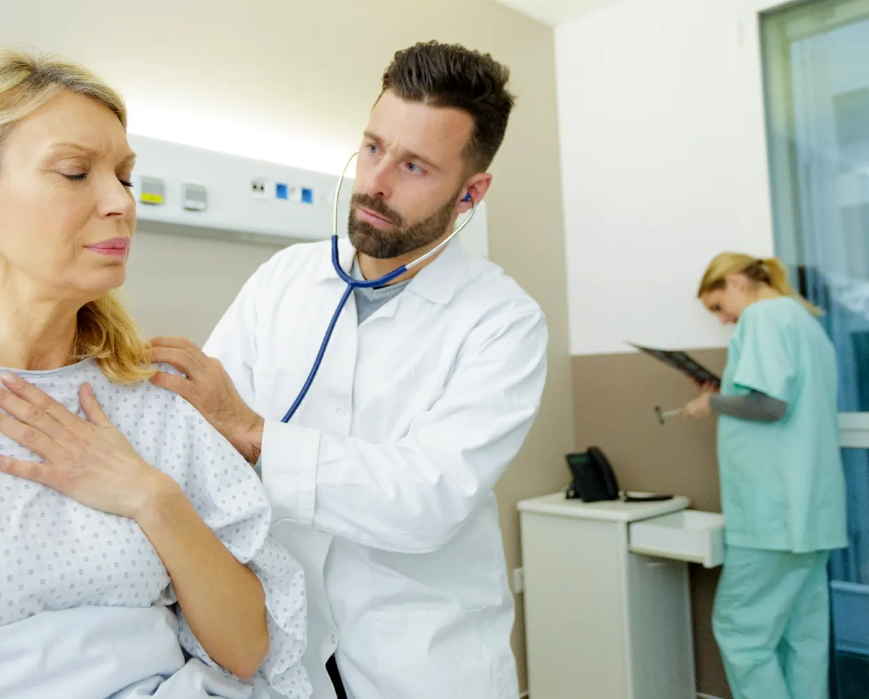 Inside a hospital patient room, a doctor uses a stethoscope to listen to a patient's heartbeat. The patient is a middle-aged woman who is grimacing, with her hand placed on her chest as if in pain.