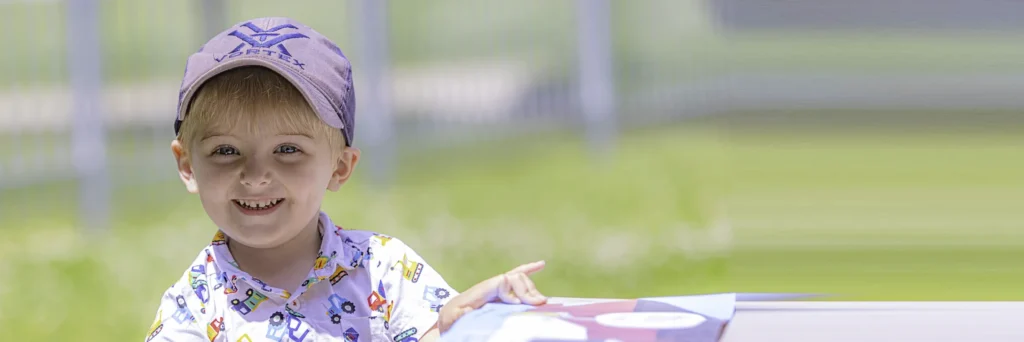 A young boy in a baseball hat sits outside on a sunny day. He's smiling and reading at a picnic table.