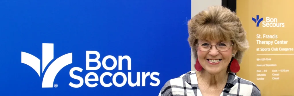 An older woman with short, blonde hair and a big smile stands in front of a blue Bon Secours sign.