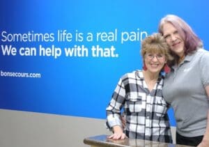 Two older woman with big smiles stand in front of a sign that says: "Sometimes life is a real pain. We can help with that."