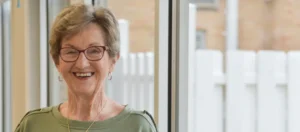 A senior woman with short, blonde hair and red-rimmed glasses smiles while standing in a well-lit hospital hallway.