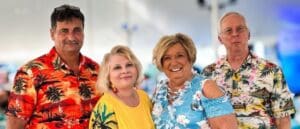 Two men and two women in floral summer shirts stand outside, under a tent. They are looking at the camera and smiling.