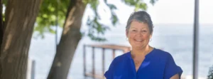 A middle-aged woman with cropped, graying hair smiles while standing outside on a summer day. Behind her is a blue, calm lake.