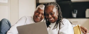 A senior couple laugh on a couch together while reviewing documents on a laptop screen.