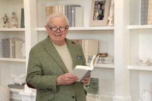 An elder gentleman in a tweed suit coat and glasses stands inside a home library. He's smiling and has an open book in his hand.