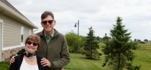 A senior couple smile while they pose for a photo together outside. They're wearing cardigans and sunglasses.