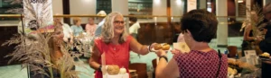 A smiling vendor serves a plate of donuts to a customer at a tasting event.