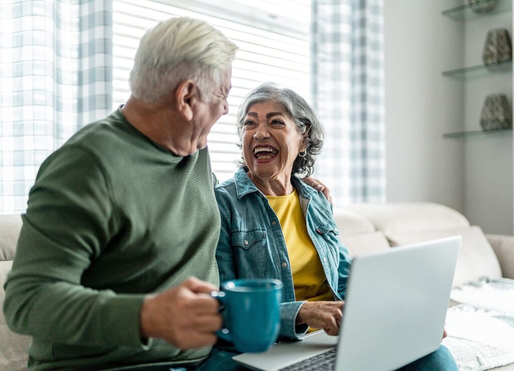 Elderly couple sitting on the couch laughing and smiling in front of their computer