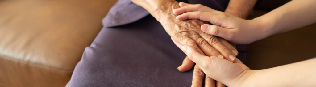 Two hands gently clasp an elderly person's hand resting on their lap