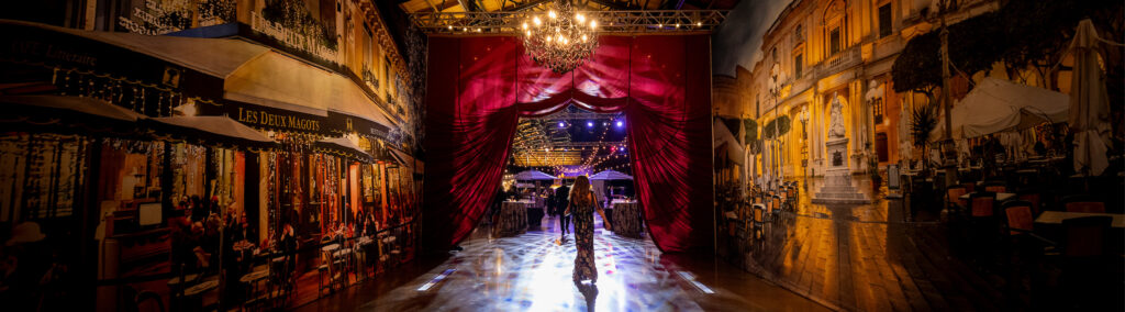 A person walks under a chandelier in a lavish room with red curtains and vibrant wall murals of outdoor Parisian cafés