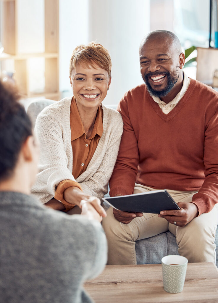 Smiling couple sits on a couch, with a woman wearing a sweater extending her hand in a handshake across a coffee table. A man holds a tablet.