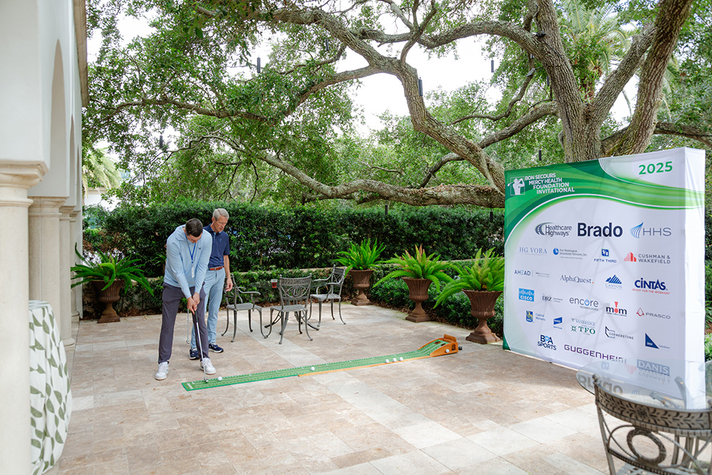 Two people play mini-golf on a patio, surrounded by lush greenery. A banner displays various sponsor logos for a 2025 charity sports event.