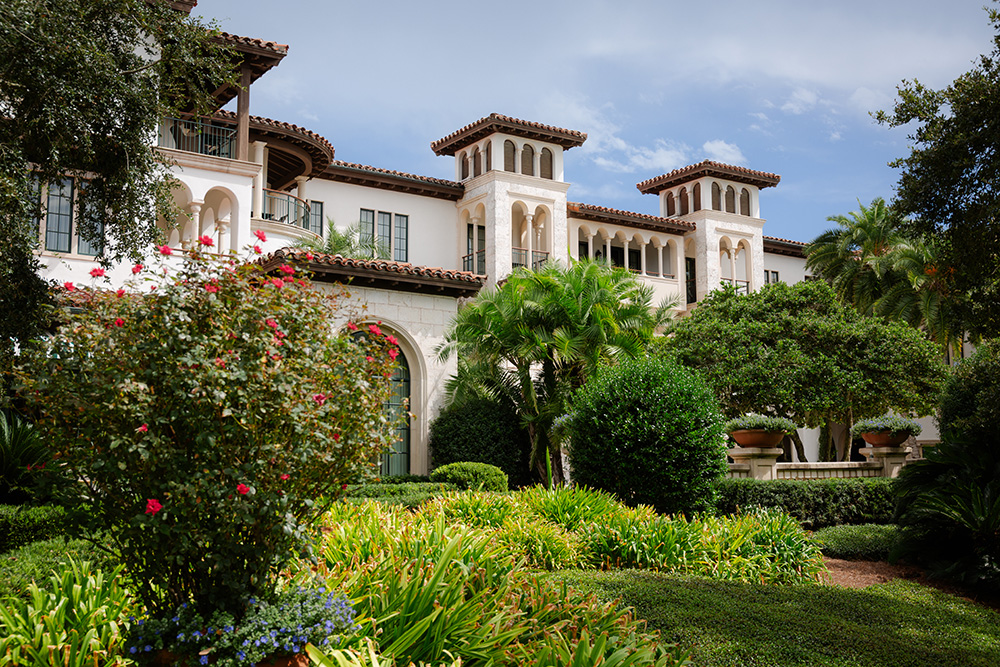A luxurious Spanish-style mansion with arched windows and terracotta roof, surrounded by lush gardens and vibrant flowers under a blue sky.