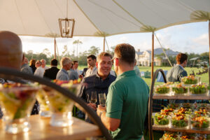 Outdoor gathering under a tent at sunset, with men chatting and holding drinks near trays of salads. A relaxed, social atmosphere is conveyed.