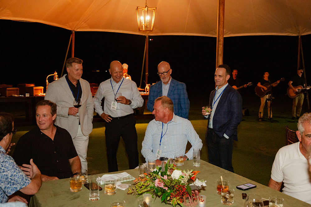 Group of men in suits enjoying a relaxed event under a tent at night. They hold drinks, engaging in conversation. A band plays in the background.
