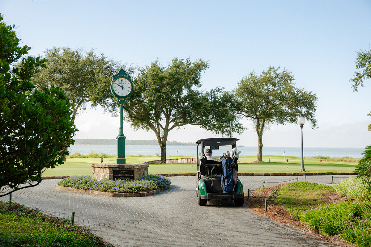 A scenic golf course view with a clock displaying 8:00, lush green grass, and a golf cart on a paved path, under clear blue skies near a calm lake.