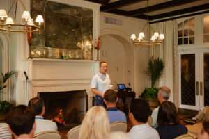 A man stands speaking to seated audience in a cozy room with elegant chandeliers, a lit fireplace, and large windows, creating an inviting atmosphere.