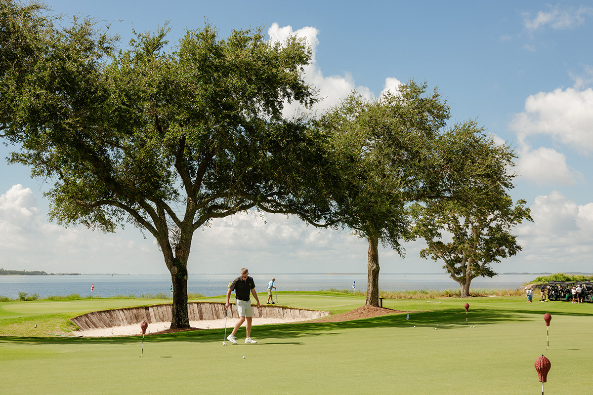 A golfer in mid-swing on a putting green beside a serene lake, framed by two large trees under a clear blue sky. Another golfer stands nearby.