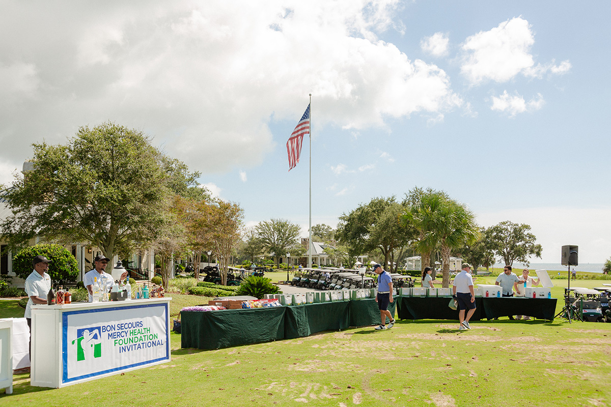 Golf course scene with tables set for the Bon Secours Mercy Health Foundation Invitational. People gather under an American flag on a sunny day.