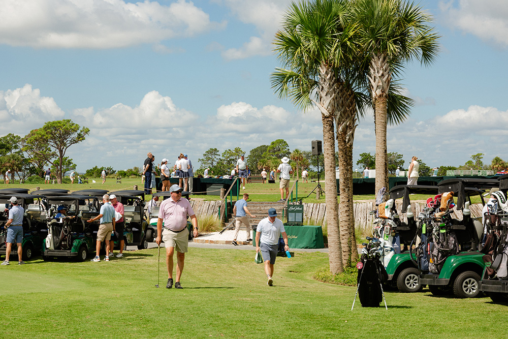 Golf course scene with people and golf carts under a sunny sky. Two tall palm trees stand prominently. The mood is lively and relaxed.