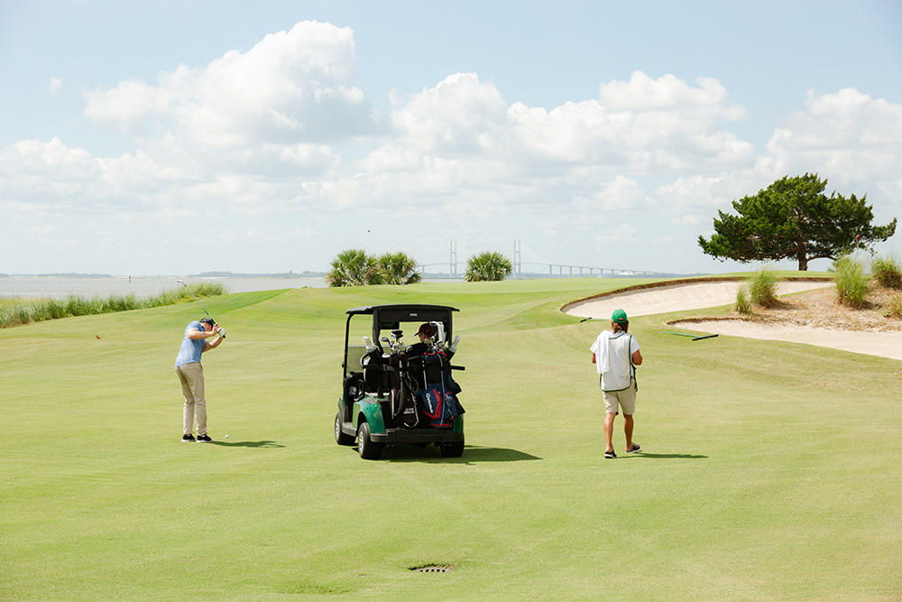 Two golfers on a sunny course with a view of a bridge in the background. One golfer swings, while the other walks near a golf cart.