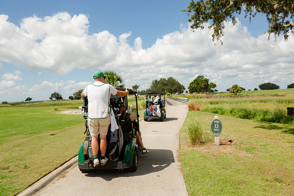 Golfers on two carts drive along a lush fairway under a partly cloudy sky, passing a sign marking hole 13. The scene feels leisurely and calm.