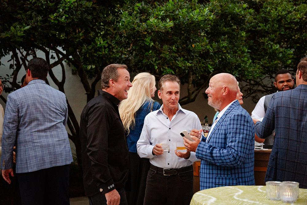 A group of people in formal attire engages in lively conversation at an outdoor event. They stand near a bar, surrounded by lush greenery, creating a warm and sociable atmosphere.