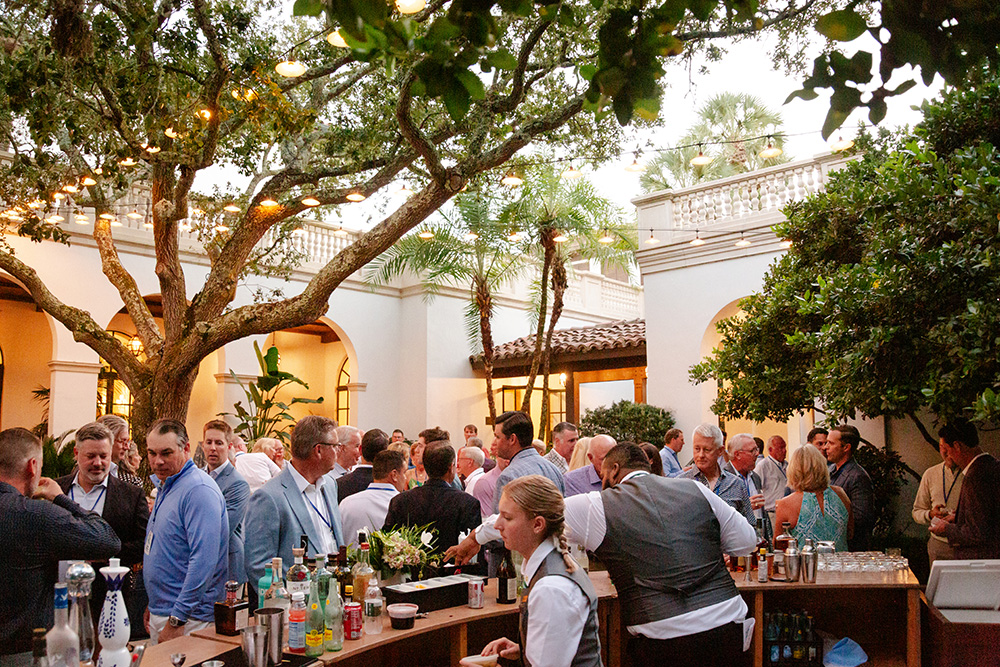 Outdoor cocktail party with attendees mingling under string-lit trees. A bartender serves drinks at a lively bar, creating an elegant and festive atmosphere.