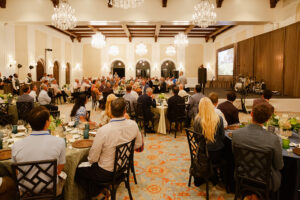 A large, elegant banquet hall with chandeliers, filled with people seated at round tables. A speaker stands on stage, creating a formal and engaged atmosphere.