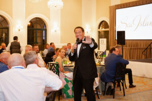 A man in a suit speaks into a microphone at a formal event with seated guests in a luxurious hall. A large screen reads "Sea Island." Joyful atmosphere.