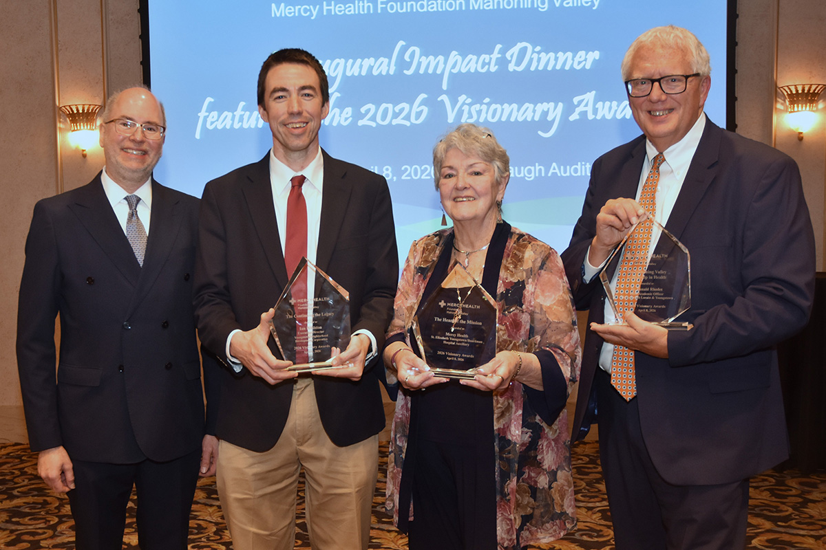 Four people stand smiling, holding glass awards at the Mercy Health Foundation event. A screen in the background reads “Inaugural Impact Dinner.”